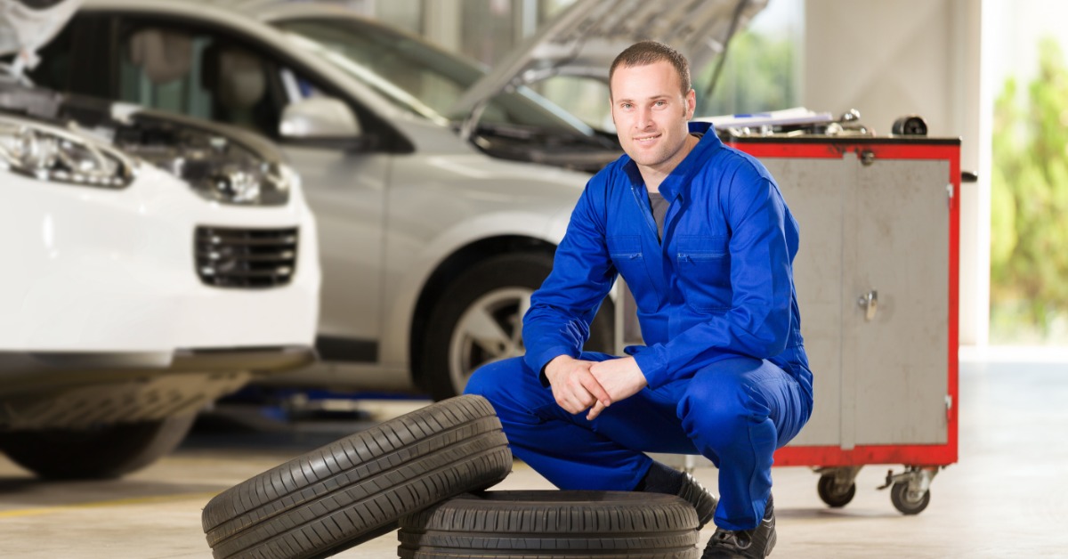 A man in a blue jumpsuit crouches in front of two car tires in front of a toolbox on wheels and two cars.