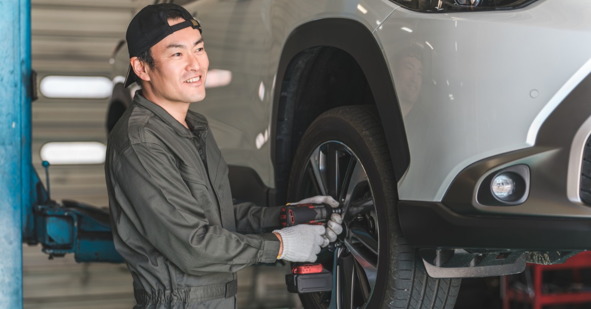 A man in a green jumpsuit and a backwards baseball hat uses a drill to tighten the lug nuts of a tire on a lift.