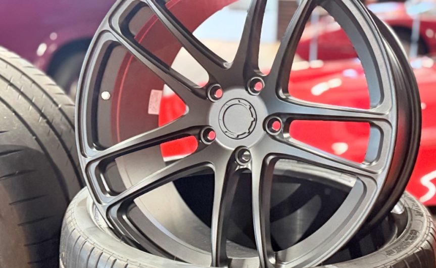 A close-up of a black empty car wheel rim standing upright on a new tire with red cars in the background.