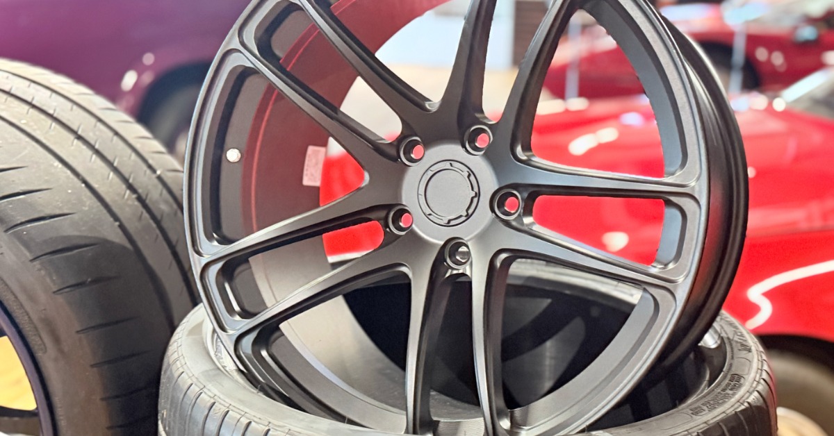 A close-up of a black empty car wheel rim standing upright on a new tire with red cars in the background.