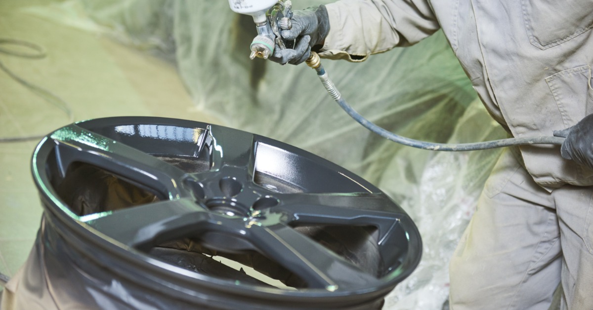 A person in a beige jumpsuit uses a paint sprayer to apply a dark grey paint coating to a car wheel.