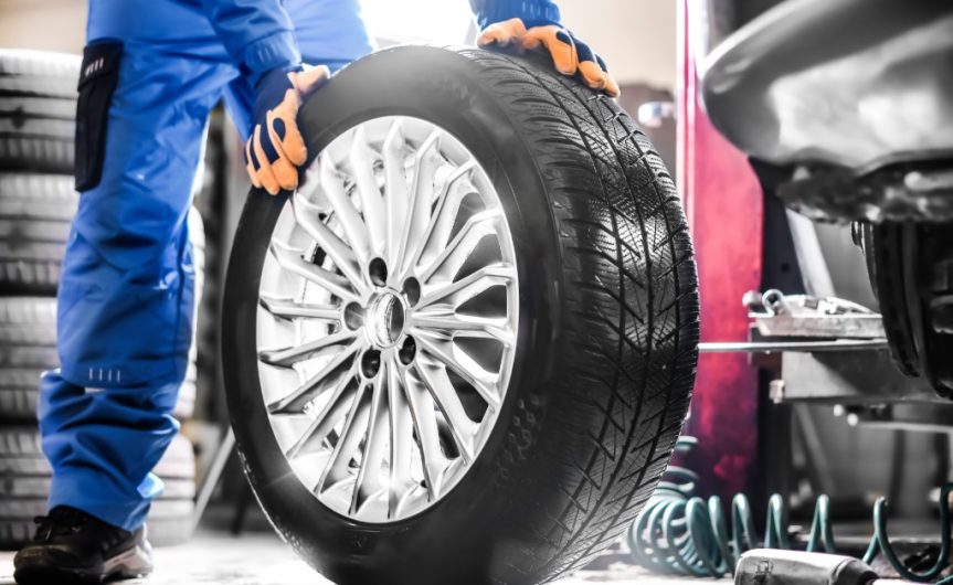 An auto mechanic in blue overalls and gloves holds a tire upright. Other auto shop equipment is nearby.
