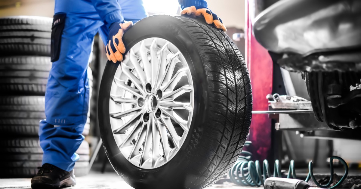 An auto mechanic in blue overalls and gloves holds a tire upright. Other auto shop equipment is nearby.
