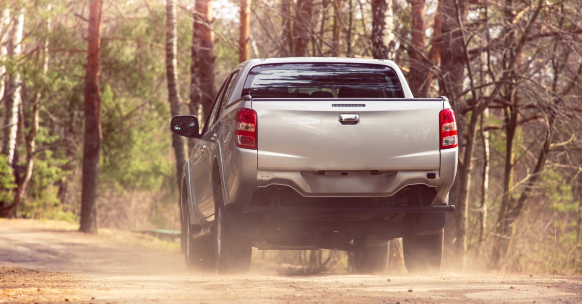 A silver pickup truck drives down a gravel road with trees on either side of the vehicle. It's dusty around the tires.