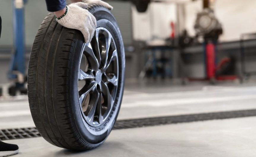 A mechanic wheeling a tire across a service shop towards an elevated car. The mechanic has white gloves on.