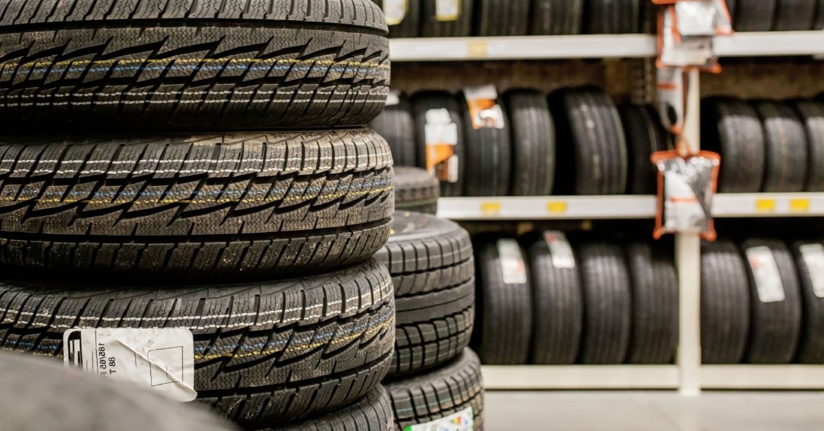 In a warehouse, a stack of car tires and wheels is set up. Behind the tires are shelves holding more tires.