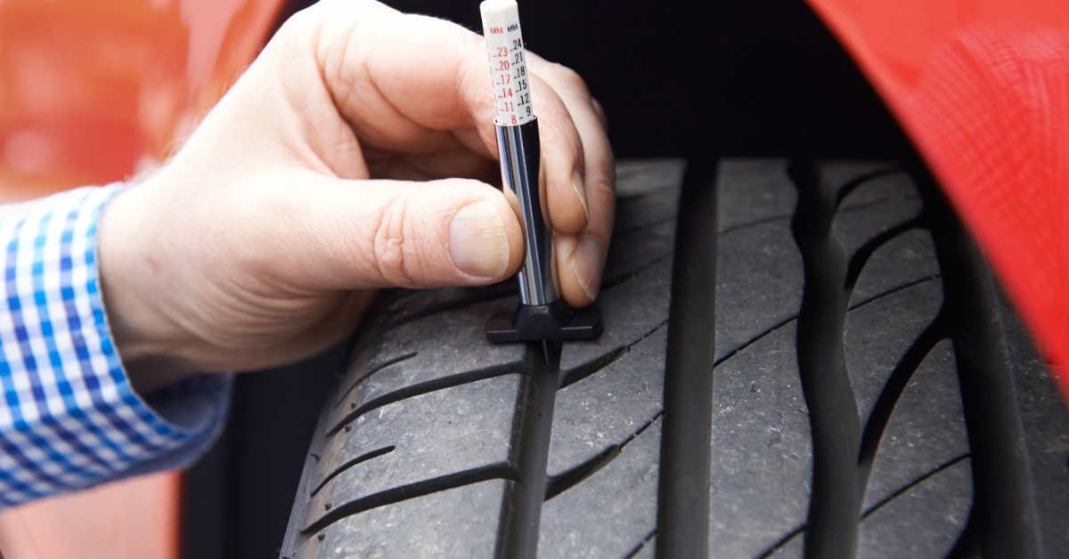 A close-up of a man's hand with a flannel shirt using a tire gauge tool to check the tire tread depth on a red car.