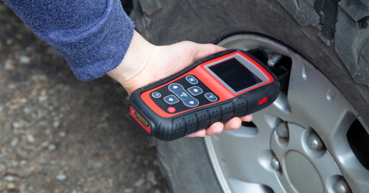 A close-up of someone holding a red TPMS pressure sensor beside the valve stem of a tire on a vehicle.