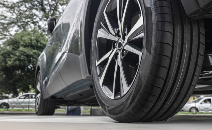 The back tire of a parked gray vehicle. The tire and vehicle are in a busy area, surrounded by other cars and trees.
