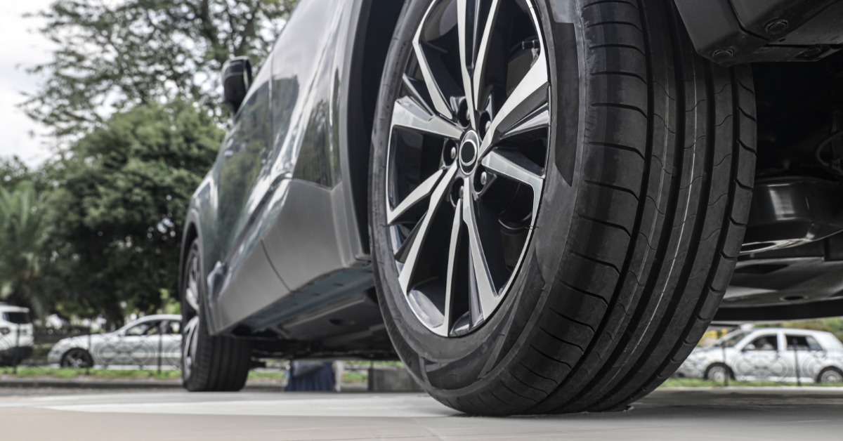 The back tire of a parked gray vehicle. The tire and vehicle are in a busy area, surrounded by other cars and trees.