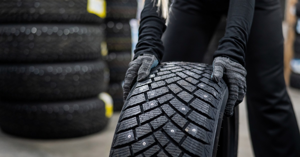 A mechanic wheeling a new studded tire out of the garage. The mechanic is wearing black overalls and gloves.