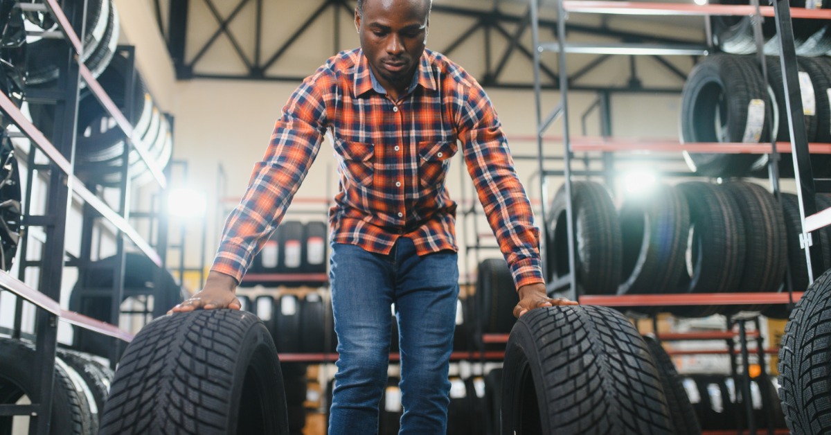 A man choosing between two tires in an automotive shop. He has both tires standing up, with a hand on each.