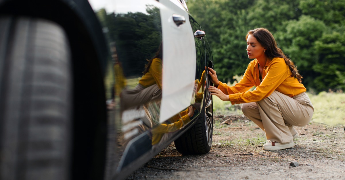 A woman in a yellow blouse and khaki pants crouching to examine the tire and wheel of a black-and-white car.