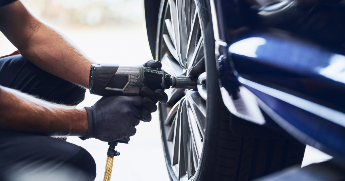 A close-up of a man crouching to use a pneumatic wrench to remove the lug nuts of a car wheel on a modern blue car.
