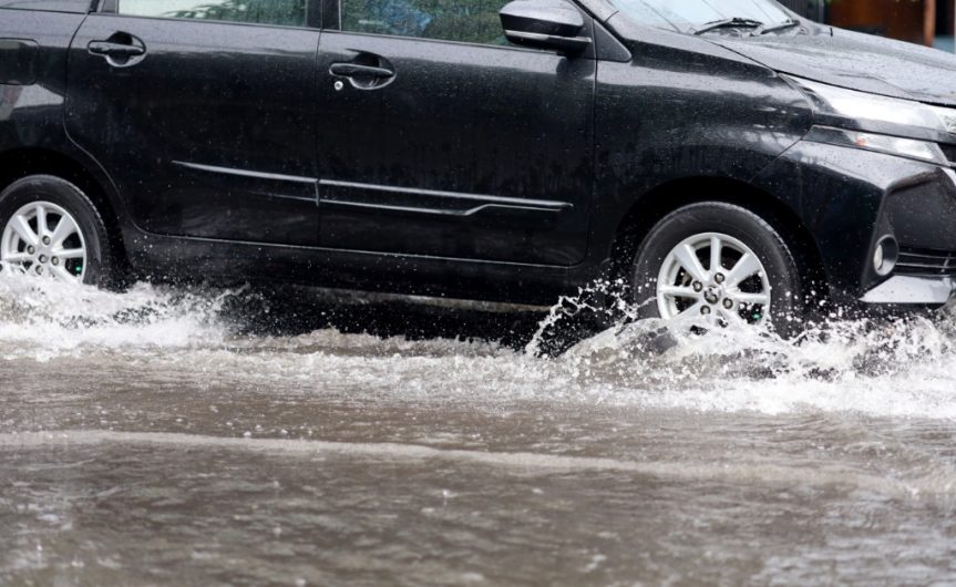A black car drives through a massive, deep puddle. Water sprays away from the tires as it ripples across the puddle.