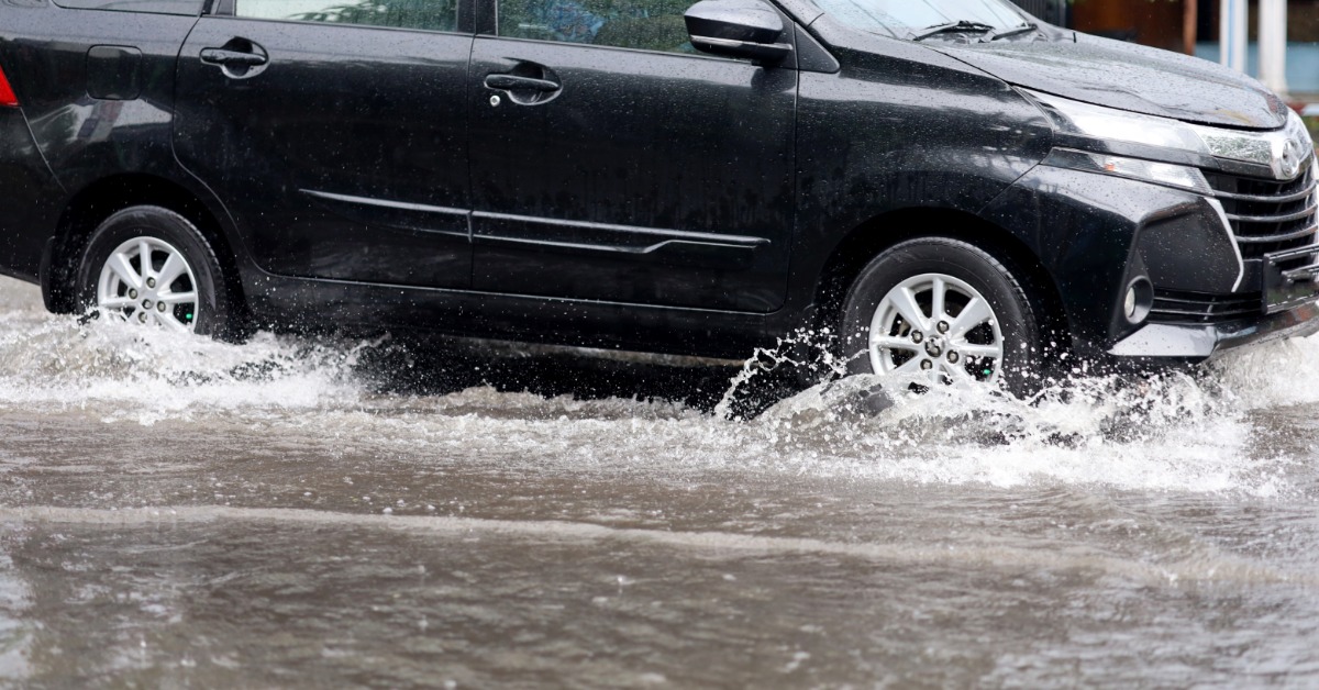 A black car drives through a massive, deep puddle. Water sprays away from the tires as it ripples across the puddle.