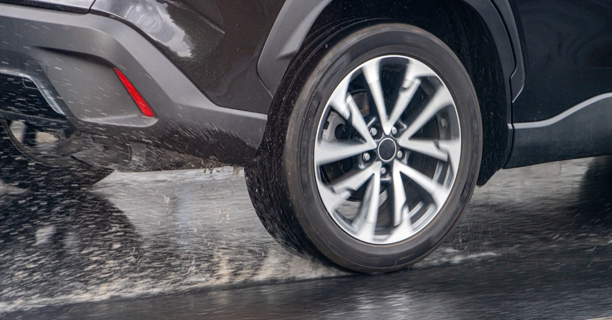 A close-up of a vehicle's tire driving over wet pavement. Water sprays around the tire with possible hydroplaning.