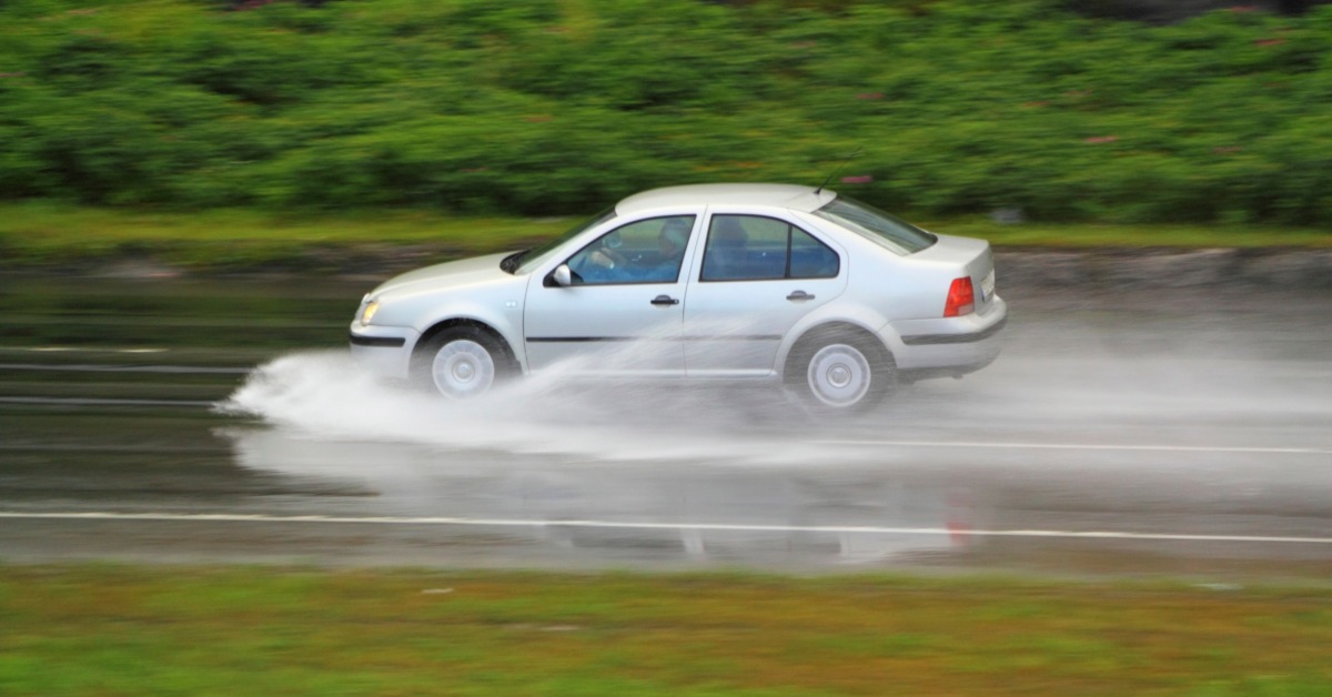 A silver sedan speeds down a wet road. Water sprays around and in front of the vehicle, with the grass surrounding the road blurred.