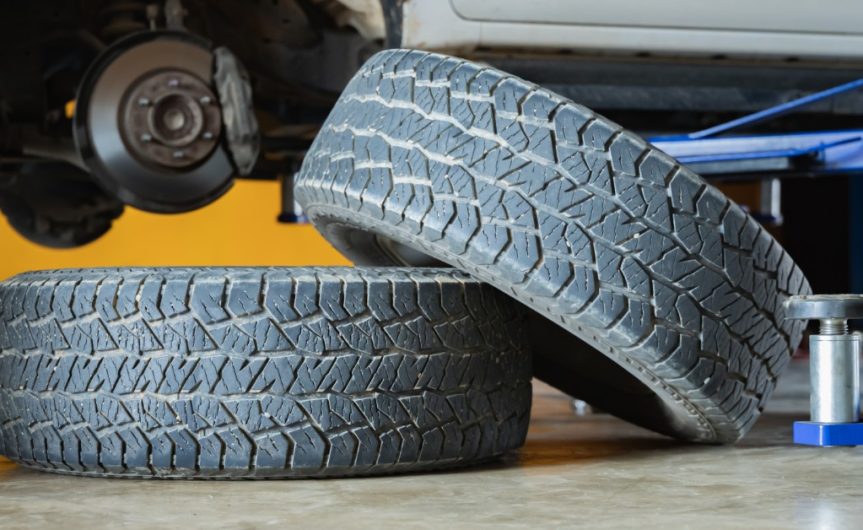Two used tires rest on a garage floor. A car is jacked up behind the tires with the back wheels missing.