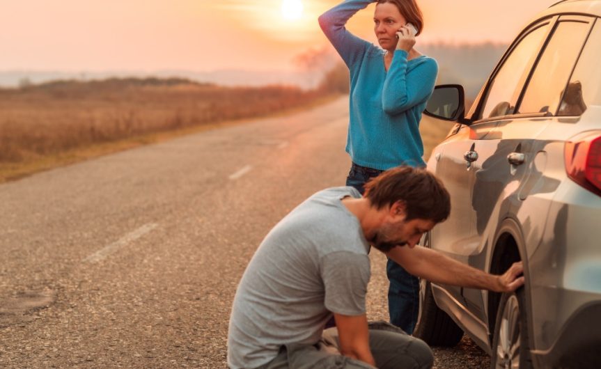 A man and a woman next to a car on the side of a road with a flat tire. The woman is talking on the phone.