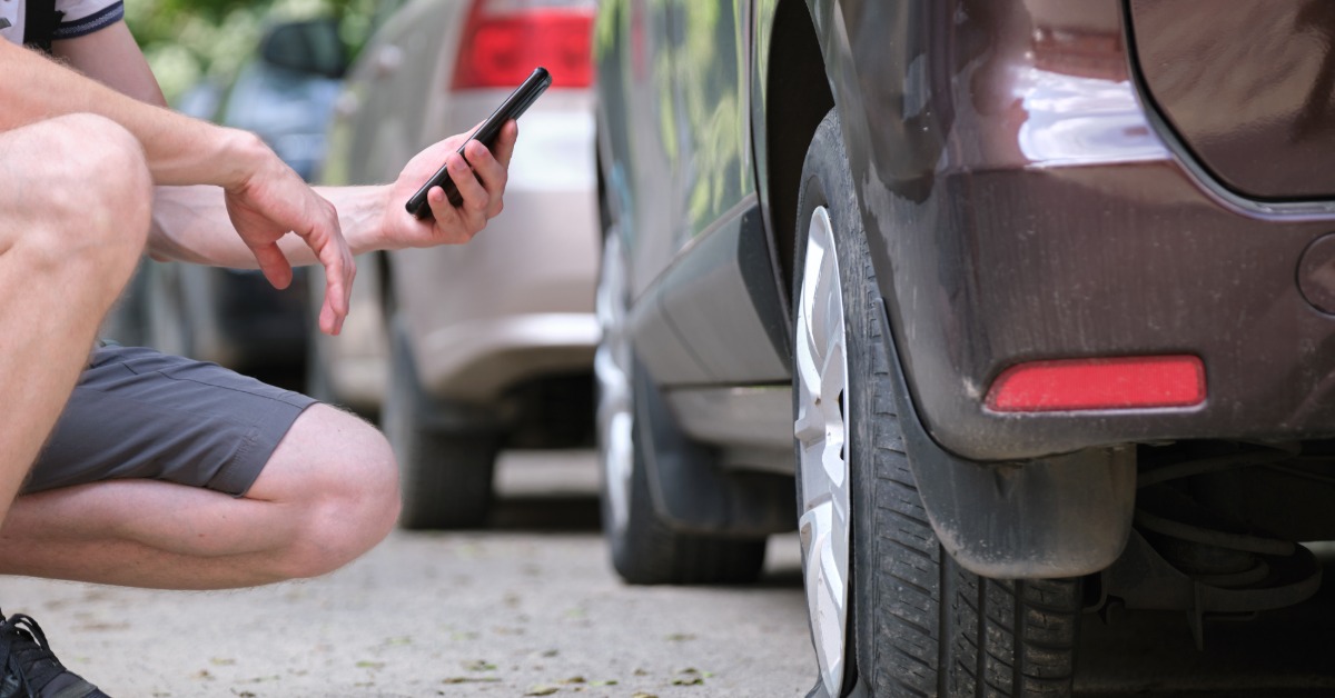 A man crouching to use his phone to take a photo of a flat tire on a black car with other cars in the background.