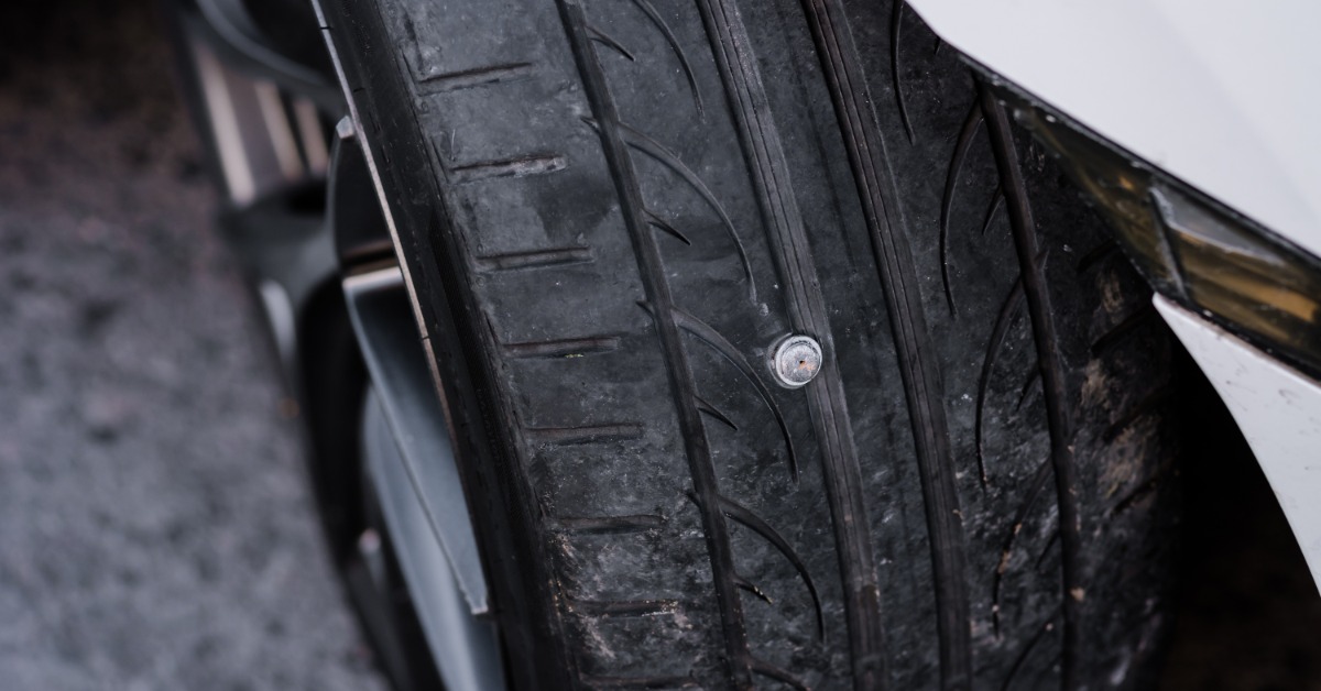 A close-up of a nail puncture on a car tire. The nail is completely embedded in the tire to only show the head.