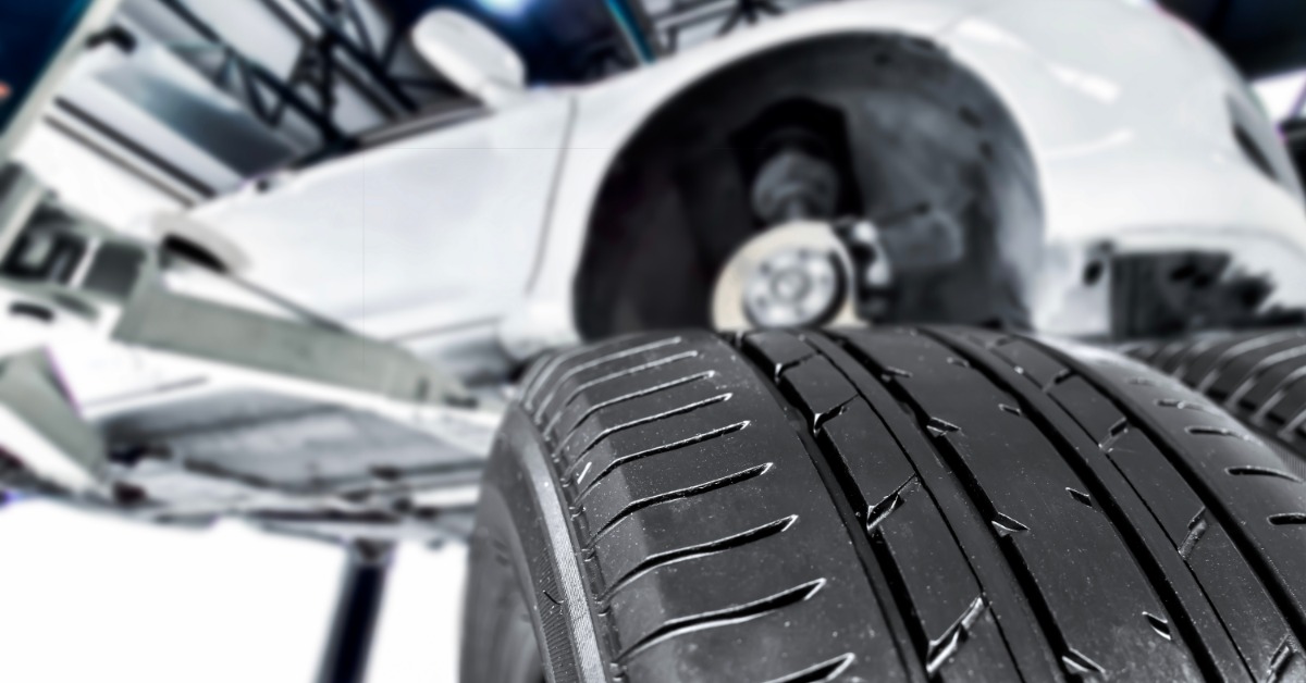 A close-up of a new tire with shiny rubber. A white vehicle is blurred and on a car lift in the background.