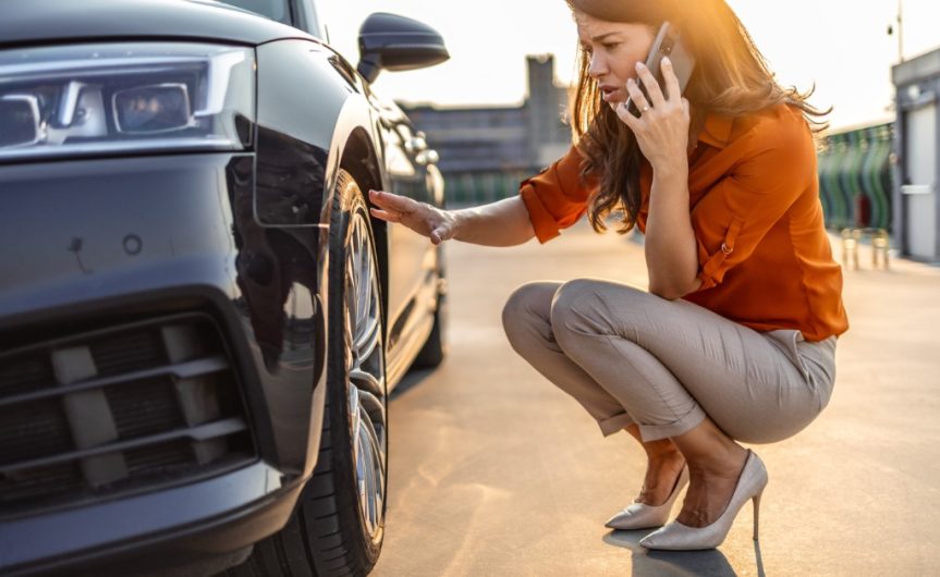 A woman in an orange blouse and high heels crouches to look at the front tire of a car while talking on the phone.