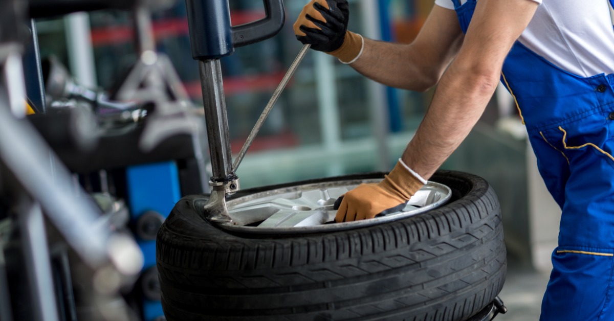 A man in blue overalls and a white t-shirt pulls the lever of a wheel balancing machine for a car wheel.