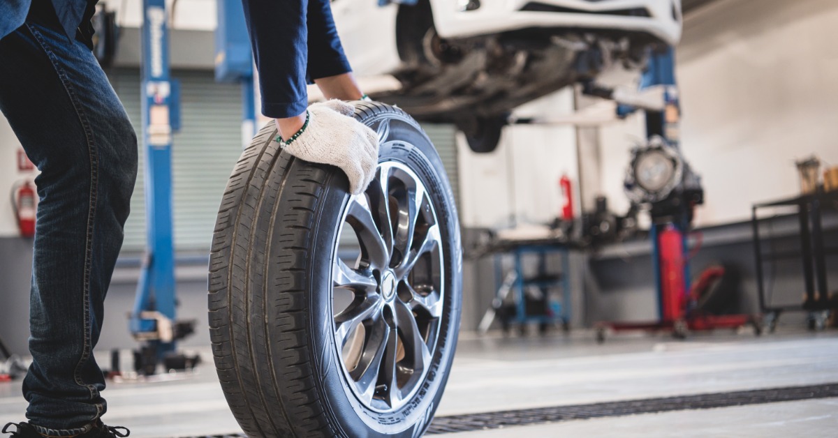 A man with white gloves crouches to wheel a car tire toward a white car elevated on a hydraulic lift.