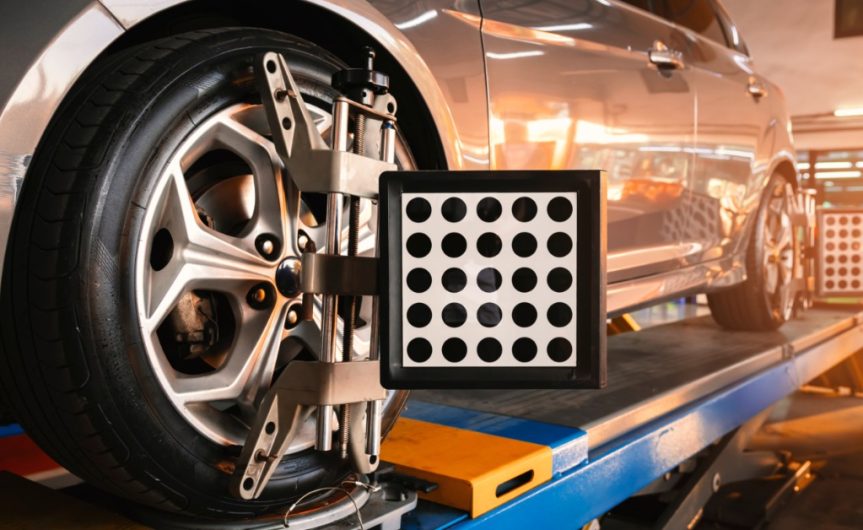 A car on a lift with computerized wheel‑alignment targets attached to the front and back tires. Lights illuminate the shop.