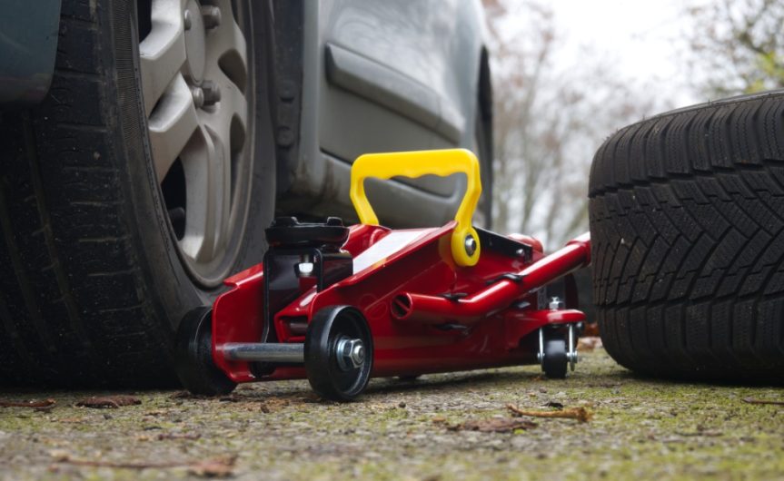 A ground-level view of a red and yellow hydraulic floor jack next to a car's tire with a spare tire next to it.
