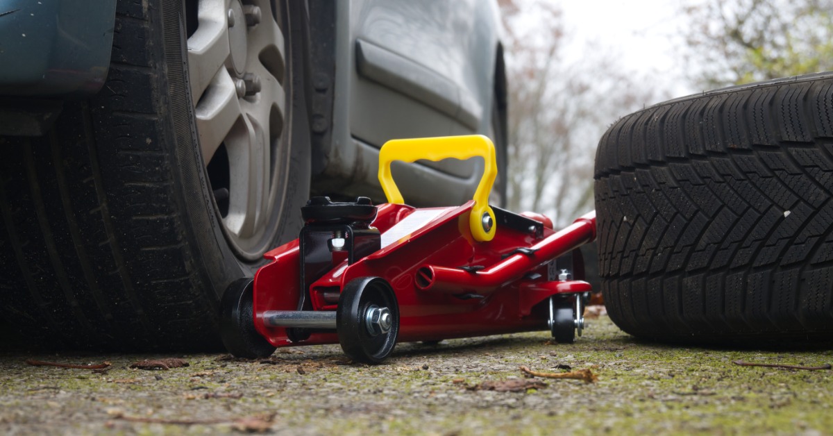 A ground-level view of a red and yellow hydraulic floor jack next to a car's tire with a spare tire next to it.