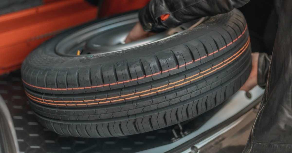 A close-up of a man in a black leather jacket removing a spare automotive tire from the trunk of a car.