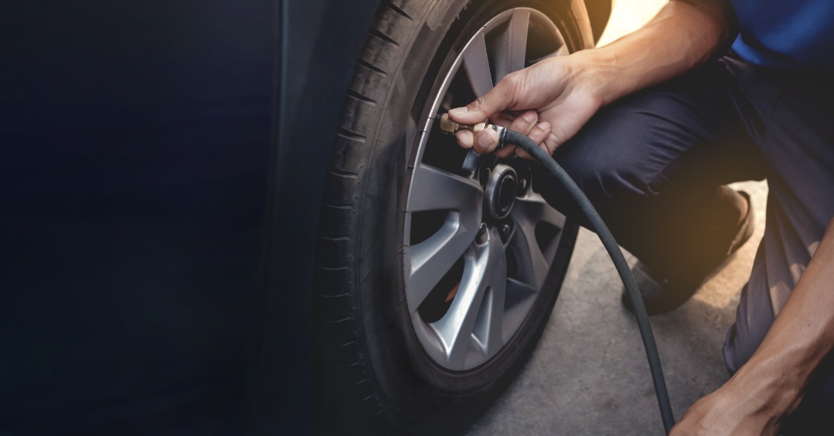 Someone kneels next to a tire on a vehicle. They hold a black air compressor hose to the tire's valve.