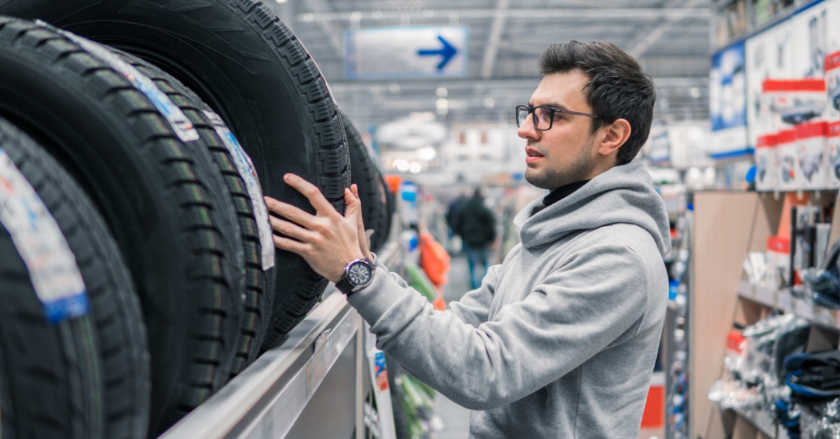 A young man in a grey hoodie with glasses is removing a new car tire from the top of a rack in a store.