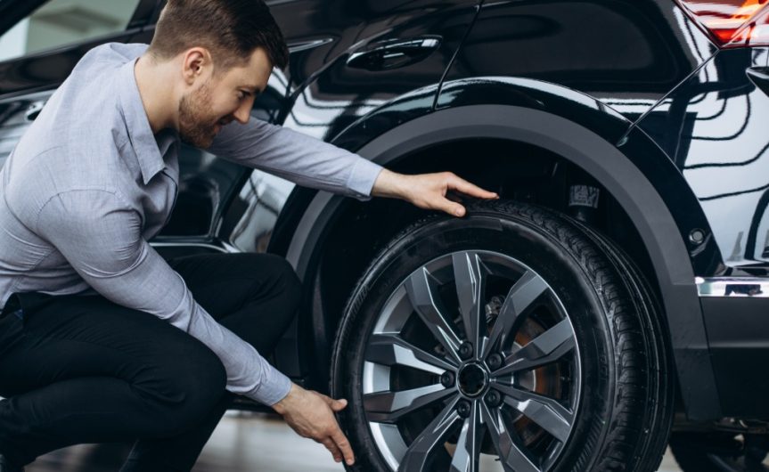 A man in a dress shirt, pants, and shoes crouches to examine the tires of a modern, black midsize SUV.