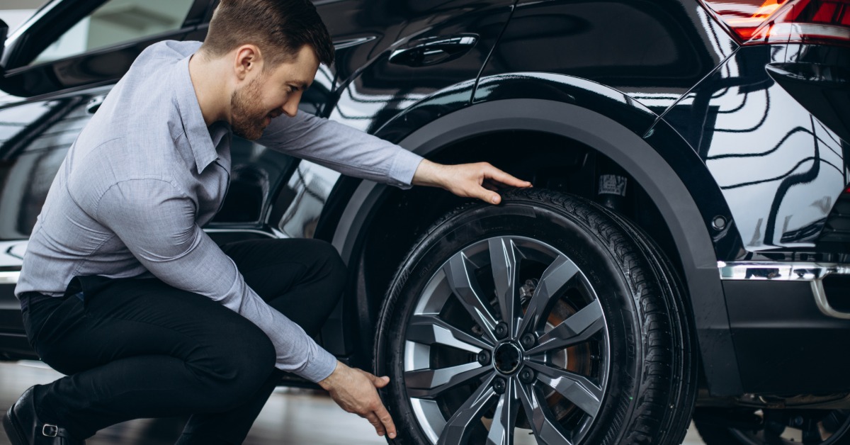 A man in a dress shirt, pants, and shoes crouches to examine the tires of a modern, black midsize SUV.