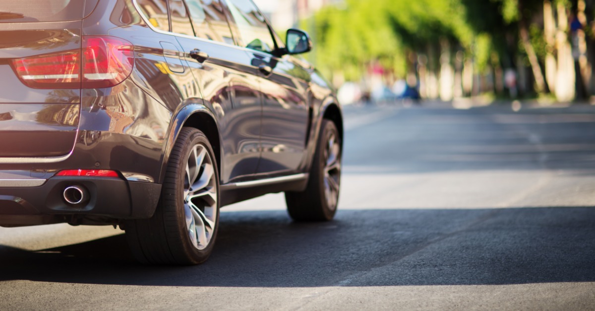 A side view of an SUV and its tires as it drives down a road. Green trees are blurred ahead of the vehicle.