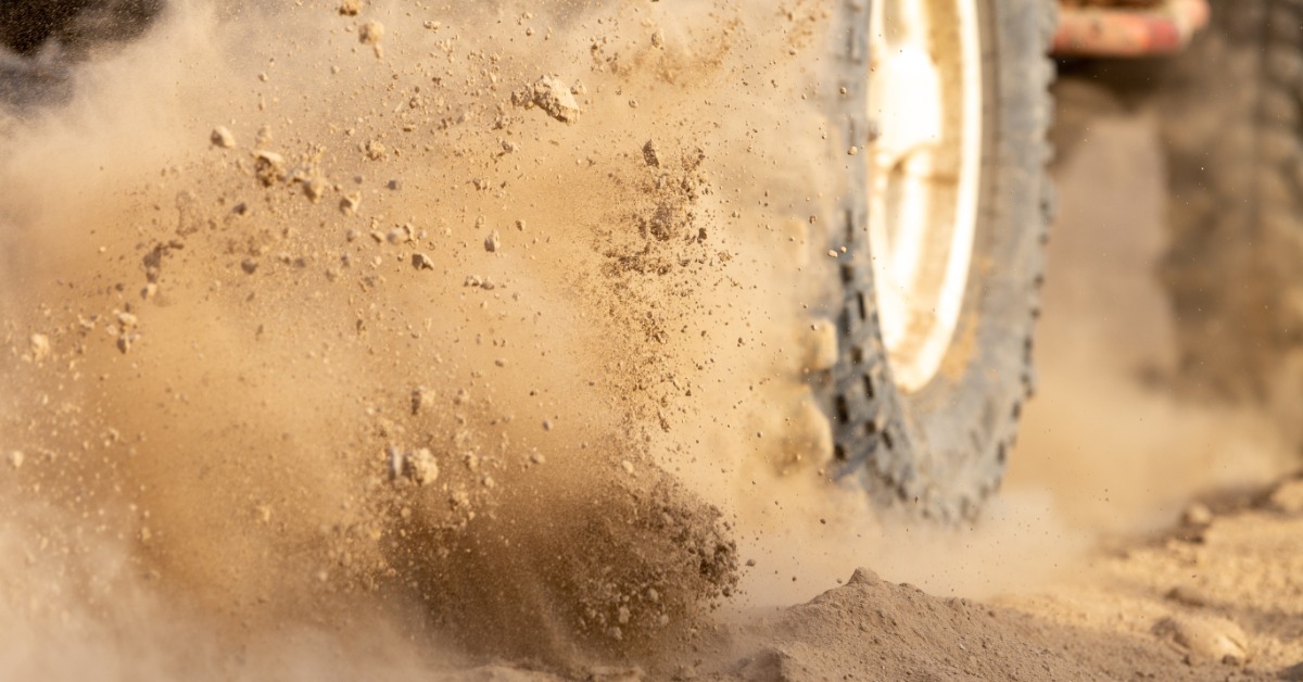 A close-up action shot of an automotive off-road tire kicking up a cloud of sand and dirt on desert terrain.