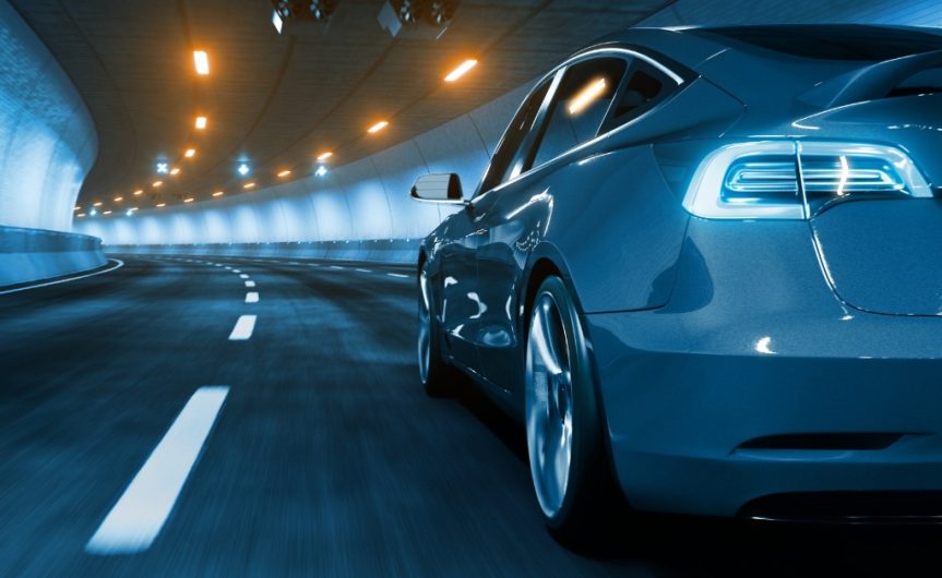A grey electric vehicle drives down a road and through a brightly lit tunnel. The white lines on the road are blurred.