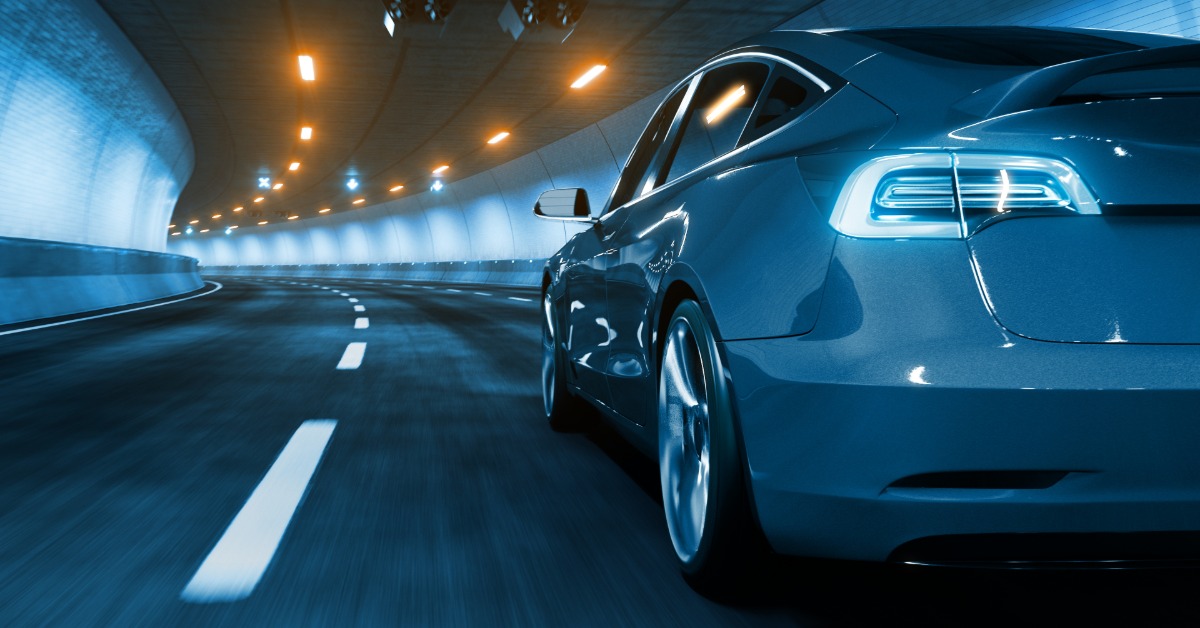 A grey electric vehicle drives down a road and through a brightly lit tunnel. The white lines on the road are blurred.