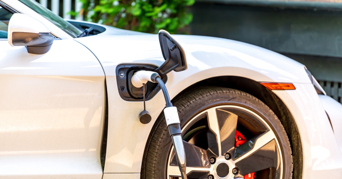 A white electric vehicle with a charging cable connected above the front passenger tire. The tire has silver rims.
