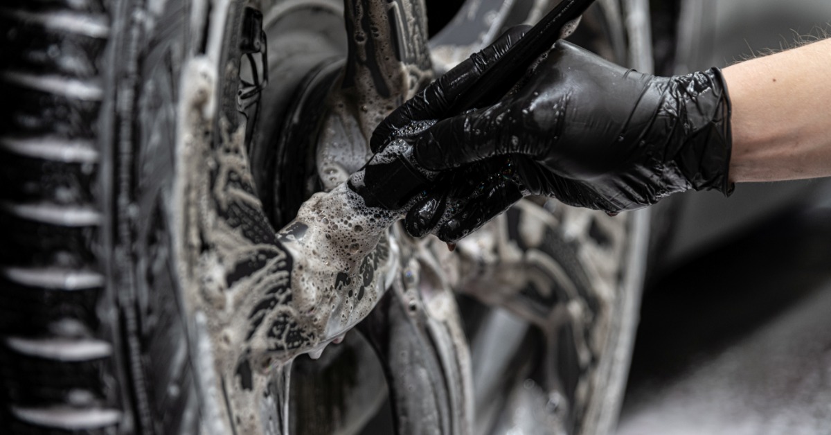 A close-up of someone wearing a black latex glove and holding a brush. They scrub the wheel of a car with soap suds everywhere.