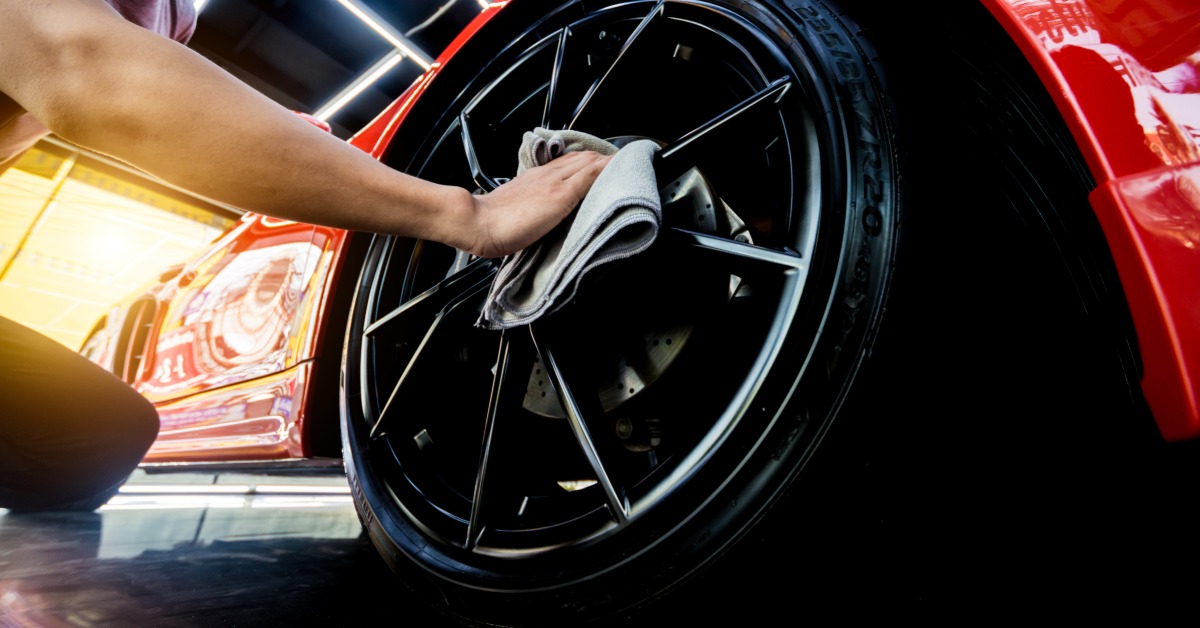 Someone kneels on the ground next to a shiny red vehicle. They use a microfiber towel to dry the black rims of the wheel.