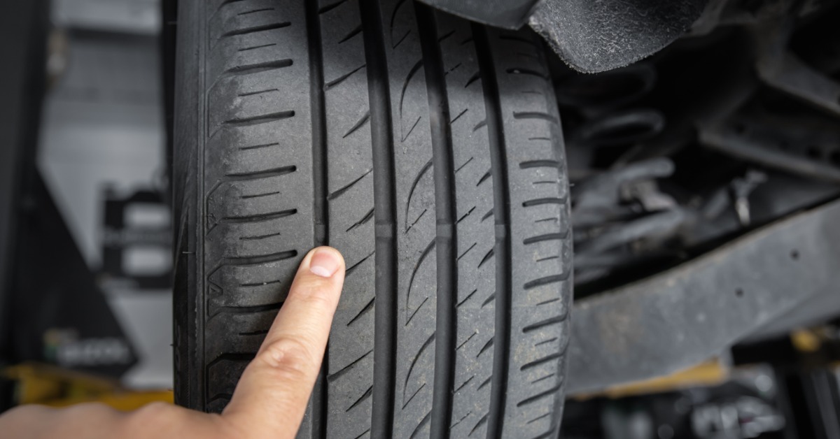 A close-up of a person's fingers pressing on the tread of a used car tire elevated on a lift in a garage.
