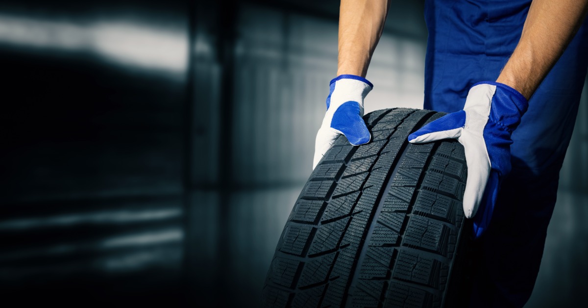 A close-up of a man with blue and white gloves and blue pants holding up an automotive tire to show its tread pattern.
