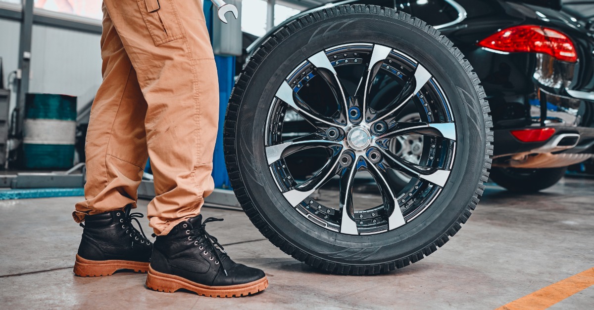A close-up of the lower body of a mechanic in an auto shop with two wrenches standing next to a tire and wheel.