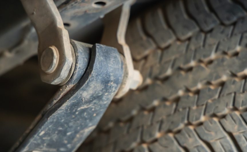 On the undercarriage of a car, the suspension of a car is dirty. The tire tread is visible behind the suspension.