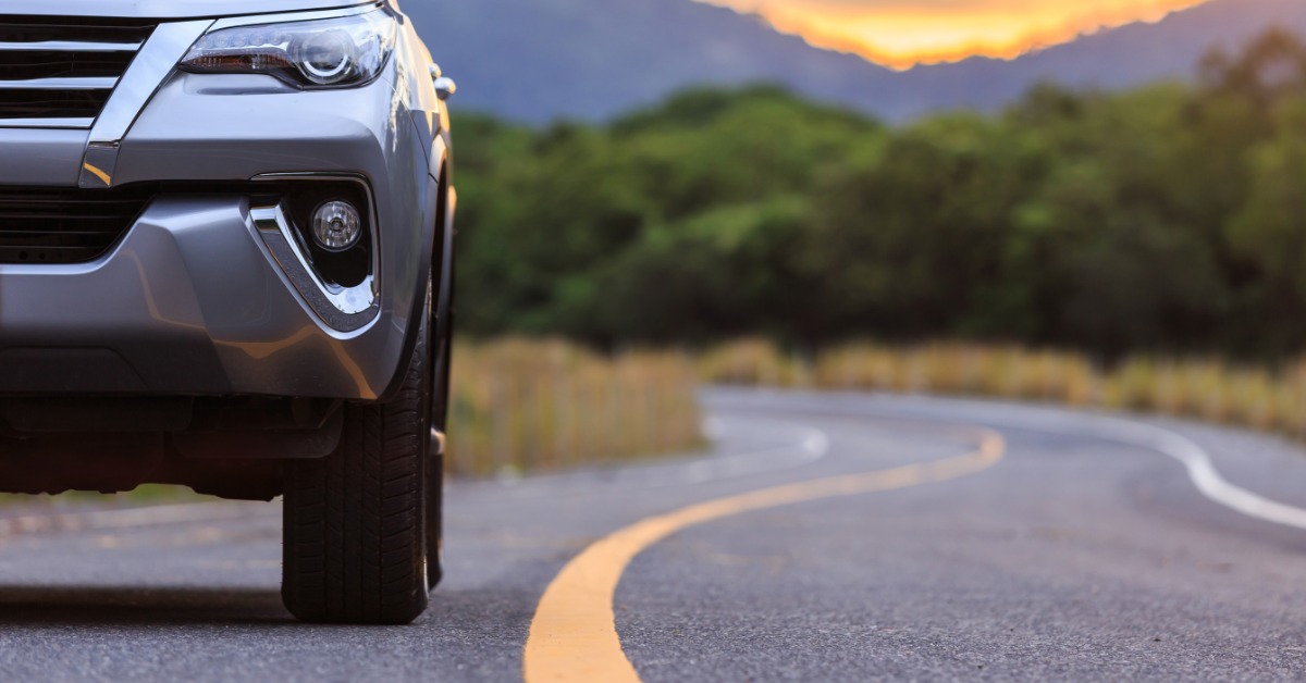 A silver SUV driving on an open road, next to a solid yellow line. There are trees along the road behind the car.